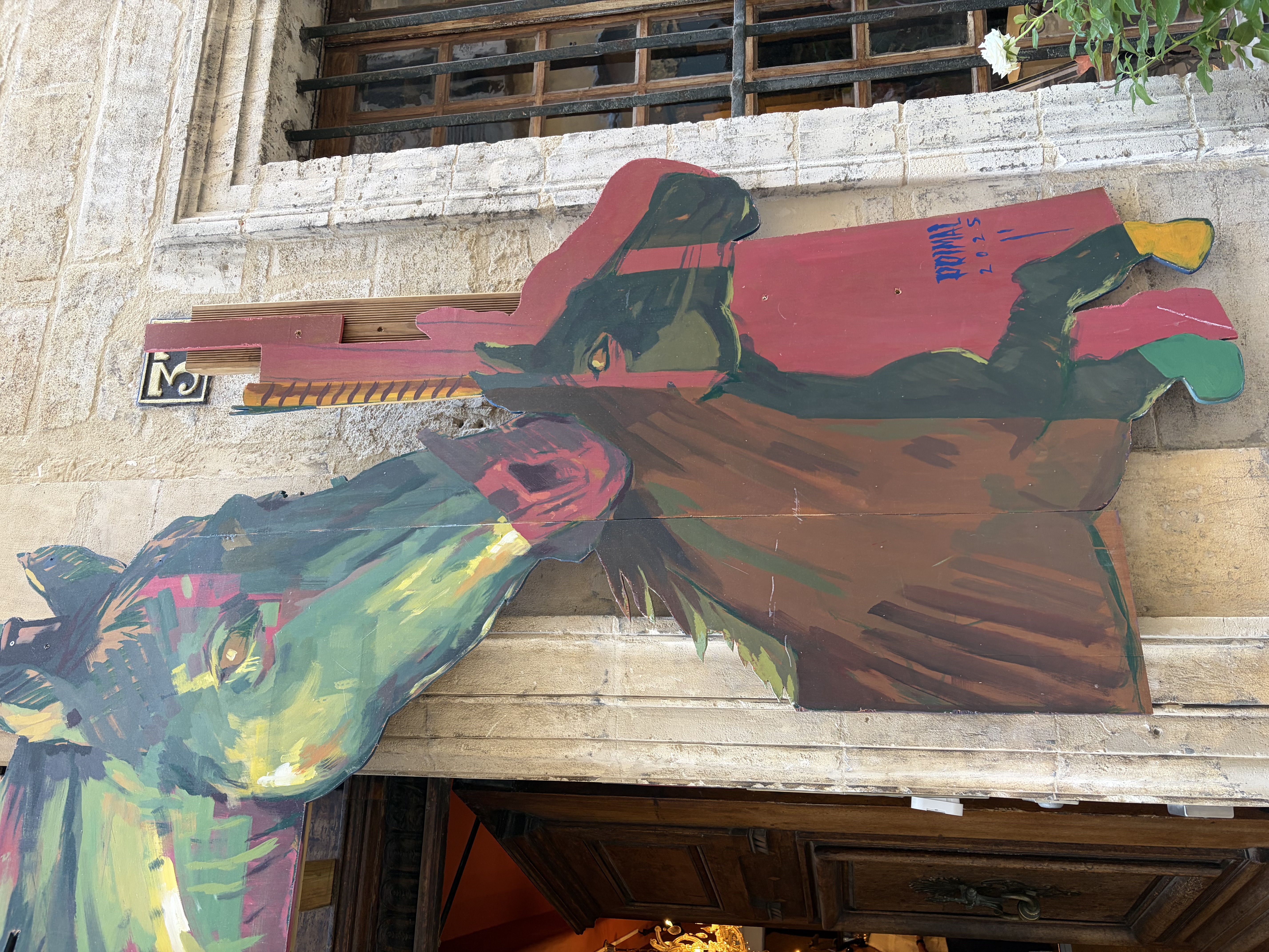 Lebensgrosse Wandmalerei einer Frau im grauen Gewand auf rauem Kalkputz, daneben Olivenzweige und ein schmiedeeisernes Schild in einer Altstadtgasse der Provence