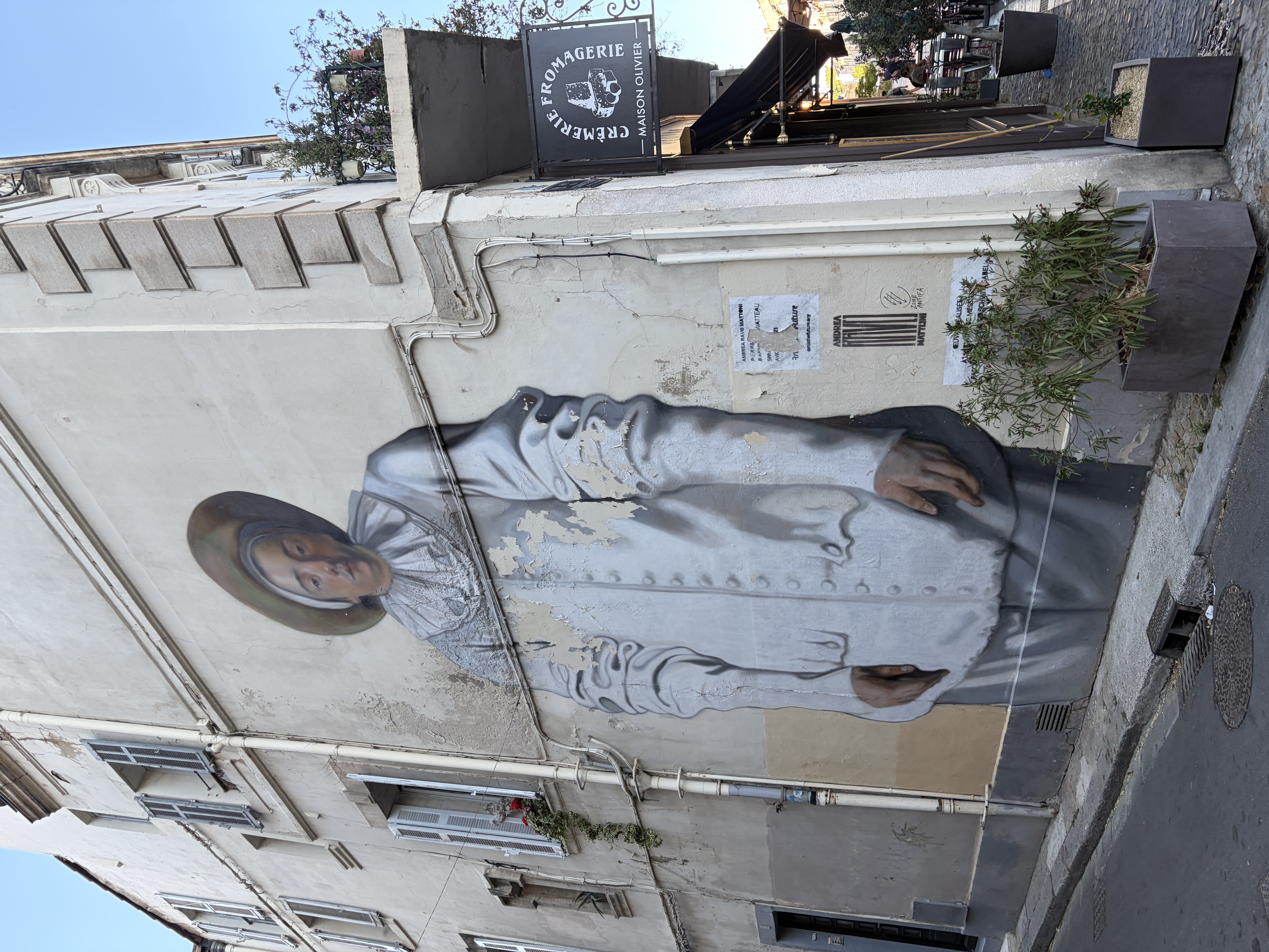 Peinture murale grandeur nature d'une femme en robe grise sur un enduit à la chaux rugueux, à côté des branches d'olivier et une enseigne en fer forgé dans une ruelle de la vieille ville en Provence