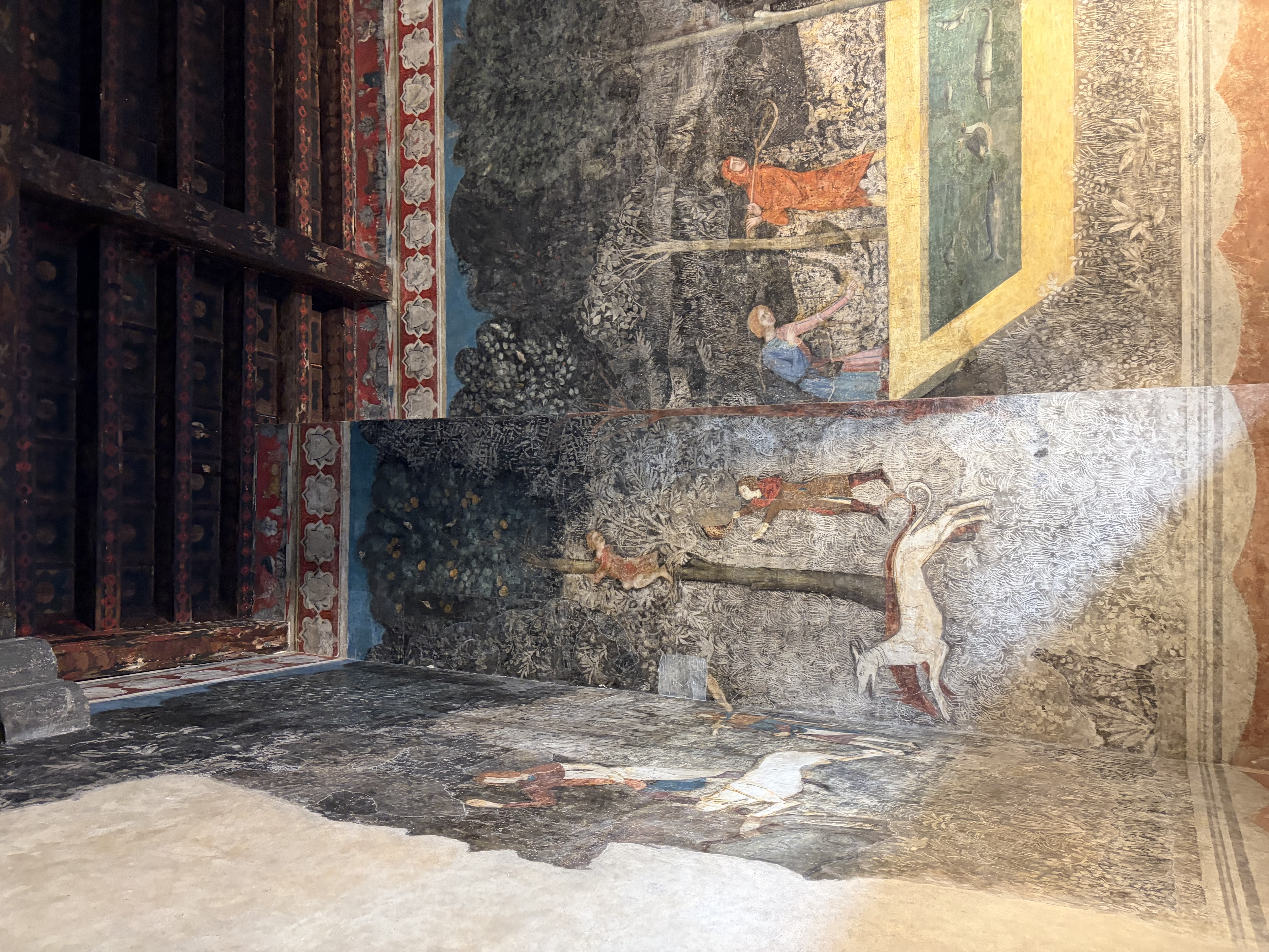 Verwitterte hellgraue Steinmauer eines provenzalischen Gebäudes mit regelmäßig angeordneten Fenstern. Davor ein akkurat gepflegter Garten mit grünen Hecken, violetten Blüten und mediterranen Pflanzen. Eine Galerie verbindet Haus und Garten.