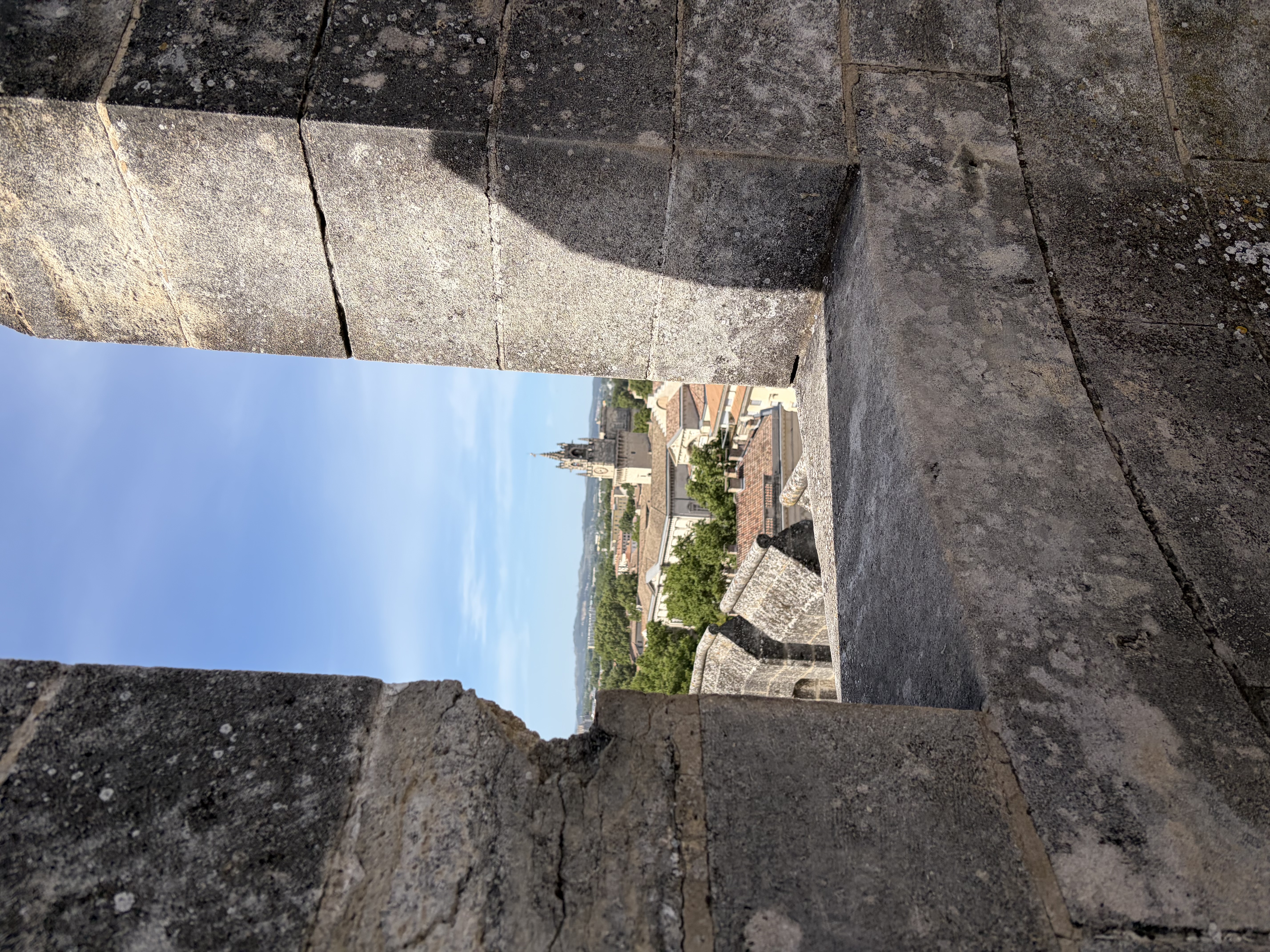 Verwitterte hellgraue Steinmauer eines provenzalischen Gebäudes mit regelmäßig angeordneten Fenstern. Davor ein akkurat gepflegter Garten mit grünen Hecken, violetten Blüten und mediterranen Pflanzen. Eine Galerie verbindet Haus und Garten.